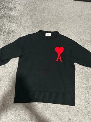 Svart stickad tröja från Ami Paris - Säljer en svart stickad tröja  Ami Paris med deras ikoniska röda hjärta och A på bröstet. Tröjan har rund hals, ribbade muddar och lång ärm. Perfekt för dig som gillar stilrena plagg med en twist. Priset kan diskuteras!