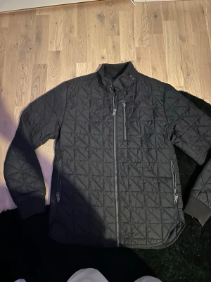 Svart quiltad jacka från Replay - Säljer en svart quiltad jacka från Replay med dragkedja framtill och tre fickor med dragkedja. Jackan har hög krage med tryckknapp och ribbade muddar vid ärmsluten. Perfekt för dig som gillar stilren och sportig look.