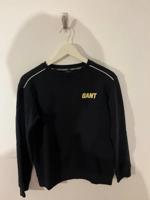 Svart sweatshirt GANT SPORT 170 cm - Mörkblå sweatshirt från GANT SPORT med stora gula och vita bokstäver på bröstet och ryggen. Klassisk rund halsringning och vita detaljer vid axlarna. Tillverkad i mjuk bomullsmix, perfekt för en sportig och avslappnad look. Använd flertal gånger men fortfarande i ett bra skick. MVH SIEO Fashion