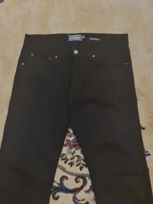 Svarta regular fit jeans från Dressmann - Säljer ett par svarta jeans från Dressmann i regular fit. Klassisk femficksmodell med raka ben och knappgylf. Jeansen har en stilren look och passar perfekt till en avslappnad stil. Materialet är slitstarkt och bekvämt. Använd bara en gång eller två