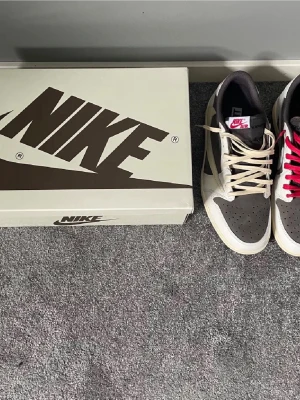 Nike Air Jordan 1 Low Travis Scott - Säljer ett par Nike Air Jordan 1 Low Travis Scott med ikonisk omvänd swoosh. Skorna har en kombination av brunt och vitt i mocka och skinn, med beige snörning och röda detaljer på hälen. Kommer med originalbox och specialdesignat rosa papper.