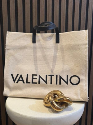 Valentino beige shoppingväska - Stilren shoppingväska från Valentino i beige bomullscanvas med svarta detaljer och stort logotryck framtill. Väskan har både handtag och avtagbar axelrem i svart skinn samt guldfärgade metalldetaljer. Perfekt för dig som vill ha en trendig och rymlig väska.