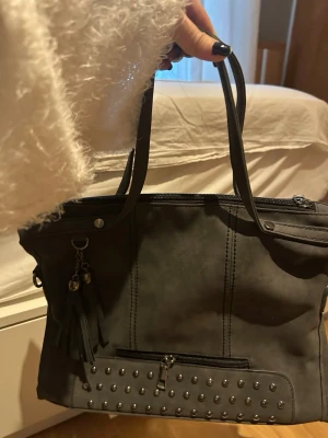 Mörkgrå mocka handväska med nitar och tofsar - Säljer en mörkgrå axelväska i syntetmaterial med coola nitar framtill och dekorativa tofsar. Väskan har både handtag och avtagbar axelrem, samt en dragkedja och en extra ficka framtill. 