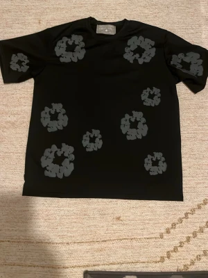 Svarta shorts från Denim Tears - Säljer ett par svarta shorts från Denim Tears med unikt blommigt mönster i mörkgrått. Shortsen har normal passform och elastisk midja med snörning för extra komfort. Perfekta för dig som vill sticka ut med en streetwear vibe.