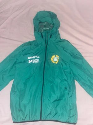 Hammarby vindjacka - Säljer en grön vindjacka från Craft med Hammarby-emblem och Intersport-logga på bröstet. Jackan har huva, hel dragkedja framtill och två sidofickor. Tillverkad i lätt polyester, perfekt för blåsiga dagar. Passar dig som gillar sportig stil.