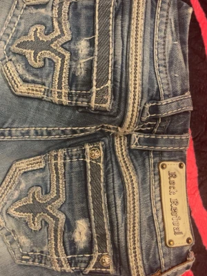 Rock Revival blå bootcut jeans - Säljer ett par blå Rock Revival jeans med slitna detaljer och kontrastsömmar. Jeansen har bootcut-modell, låg midja och coola broderade fickor. Materialet är klassisk denim i bomull och de har en snygg tvättad look med slitningar på benen.
