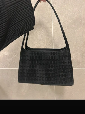 Svart axelväska från Fendi - Snygg svart axelväska med klassiskt FF-mönster över hela väskan. Väskan har en stilren rektangulär form, långa handtag och dragkedja upptill samt en extra ficka med dragkedja på baksidan. Mått: 29 x 20 x 9 cm