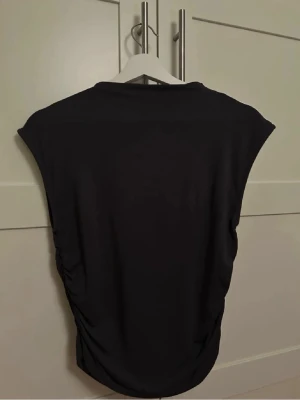 Croppad topp med rynkade sidor - Snygg svart croppad topp med höga axlar och rynkade sidor. Toppen är ärmlös och har en tight passform som framhäver figuren. Färgen är mörkgrå.