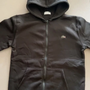 Lacoste zip hoodie - Svart hoodie från Lacoste med klassisk krokodillogga på bröstet. Tröjan har huva, hel dragkedja framtill och två stora fickor. Perfekt för en chill och stilren look.