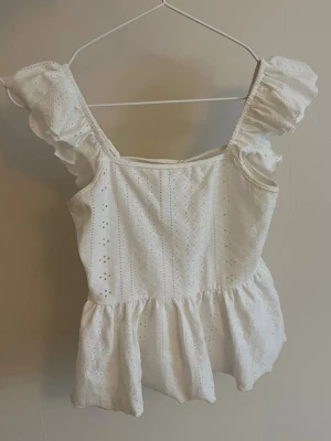 Vit blus med volang och knyt - Supersöt vit blus i bomull med broderade detaljer och volang vid axlarna. Toppen har fyrkantig ringning, korta puffärmar och en volang nertill. Baksidan har öppen rygg med knytband.