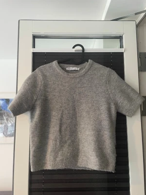 Grå stickad kortärmad tröja Zara - Mysig grå stickad tröja från Zara med korta ärmar och rund hals. Tröjan har ribbade muddar vid ärmslut och nederkant, vilket ger en clean och enkel look. Perfekt att styla med jeans eller kjol för en chill vibe.