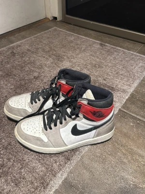 Nike Air Jordan 1 Retro High OG - Säljer ett par Nike Air Jordan 1 Retro High OG med svart swoosh, vit läderbas, grå mockadetaljer och röd krage. Klassisk high-top siluett med svarta skosnören och perforerad tåbox. Perfekta för dig som vill ha en ikonisk sneaker med streetwear-vibe.EJ BOX