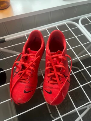 Röda Nike fotbollsskor med dobbar - Säljer ett par röda Nike fotbollsskor med svarta detaljer och klassisk swoosh på sidan. Skorna har snörning och lite slitna dobbar tyvärr. De är nog mest passande för dom nya spelarna som vill ha ett par första och kanske bara testa på sporten lite!
