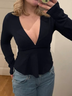 Marinblå långärmad topp med v-ringning - Säljer en marinblå långärmad topp med djup v-ringning och lätt utsvängd nederdel med knytning bak till. Toppen är i ett ribbat material som ger en snygg struktur och sitter tajt över överkroppen. Perfekt att styla med jeans eller kjol. Passar s/m