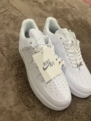 vita sneakers - Klassiska Air Force 1 sneakers i helvitt skinn med perforerad tå, vit sula och vita skosnören. Ikonisk modell med låg profil och diskret Nike-logga på sidan. Perfekta för dig som vill ha en clean och tidlös look.
