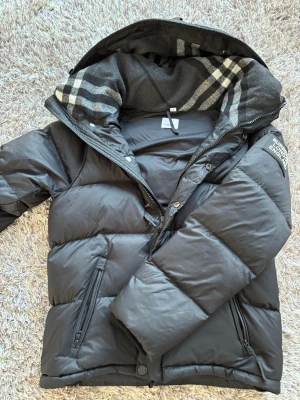 Svart pufferjacka från Burberry - Säljer en svart pufferjacka från Burberry med klassiskt rutigt foder i huvan. Jackan har dragkedja, tryckknappar och två snedställda fickor med dragkedja. Stor logopatch på ärmen och justerbar dragsko nedtill. Perfekt för kalla dagar. Man kan även ta av armarna på den så det blir en väst ✅Inga skador finns!