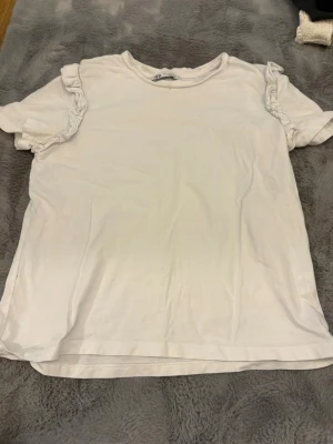 Vit t-shirt med volang från Zara - Vit t-shirt från Zara i mjuk bomull med korta ärmar och volangdetaljer längs axlarna. Enkel och clean design med rund halsringning. Perfekt för dig som gillar en söt twist på basic-plagg.