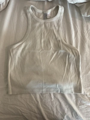 Vit ribbad linnetopp från H&M - Säljer en enkel och fräsch vit ribbad linnetopp från H&M Basic. Toppen är ärmlös med hög halsringning och croppad passform. Perfekt till sommaren och lätt att matcha med allt. Materialet är mjukt och stretchigt för en bekväm känsla.