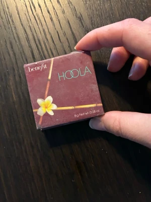Benefit Hoola Bronzer 8g - Säljer en klassisk Hoola bronzer från Benefit i originalkartong. Färgen är en matt, varm brun ton som ger en naturlig solkysst look. Kommer i en söt fyrkantig ask med spegel i locket och tropisk design. Perfekt för contouring eller att värma upp ansiktet. 8 g. Knappt använd. 