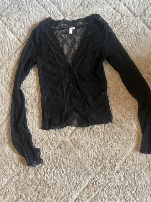Svart spetsblus med knappar, S - Säljer en svart, transparent spetsblus med lång ärm och v-ringning. Blusen har fina spetsdetaljer, knappar framtill och är perfekt för att lyfta en outfit. Snygg att styla med linne under eller som den är.