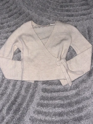 Beige omlottkofta från Gina Tricot - Supersnygg beige omlottkofta från Gina Tricot i mjukt stickat material. Koftan har långa ärmar, ribbade muddar och knyt i sidan för en extra fin detalj. Perfekt att slänga på sig under kyliga dagar!