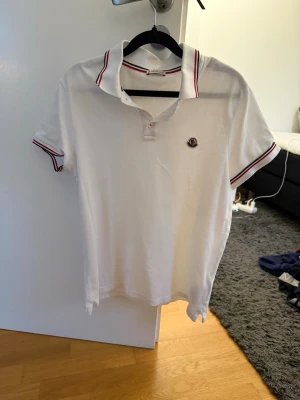 Vit Moncler pikétröja med ränder - Klassisk vit pikétröja från Moncler med korta ärmar och krage. Tröjan har röda och marinblå ränder på krage och ärmslut samt Moncler-logga på bröstet. Tillverkad i bomull för en skön och luftig känsla.