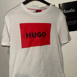 Vit t-shirt med röd HUGO-logga - Snygg vit t-shirt från HUGO med stor röd ruta och svart logga på bröstet. Klassisk passform och rund halsringning. Tillverkad i mjuk bomull som känns skön mot huden. Perfekt för dig som gillar stilrena och trendiga plagg.