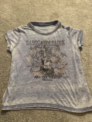 Blå Zadig & Voltaire t-shirt med tryck - Säljer en ljusblå t-shirt från Zadig & Voltaire med coolt tryck av elgitarr och blommor samt texten 'ART IS TRUTH' på framsidan. T-shirten har korta ärmar och rund hals. Perfekt för dig som gillar rockig stil och snygga detaljer.