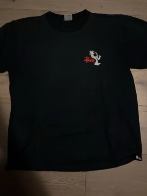 Svart Stüssy t-shirt med tryck - Svart t-shirt från Stüssy med coolt vitt tryck av en saxofonspelare och röd Stüssy-logga på bröstet och ryggen. Klassisk passform och rund hals. Perfekt för dig som gillar streetwear och vill sticka ut med en unik design.