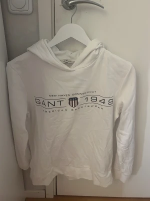 Vit hoodie från Gant med tryck - Snygg vit hoodie från Gant med svart tryck framtill där det står 'GANT 1949' och 'American Sportswear'. Är i väldigt bra skick då jag andvänt den fåtal gånger 