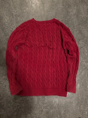 Röd kabelstickad tröja från Bison - Säljer en röd kabelstickad tröja från Bison med rund halsringning och ribbade muddar vid ärmslut och nederkant. Tröjan har ett klassiskt flätmönster och är perfekt för dig som gillar en tidlös och snygg stil. Materialet känns mjukt och bekvämt.