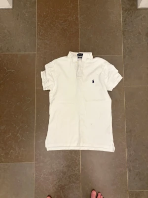 Vit pikétröja från Polo Ralph Lauren - Klassisk vit pikétröja från Polo Ralph Lauren med blå broderad logga på bröstet. Slim fit-modell med korta ärmar, krage och knappar vid halsen. Perfekt för en clean och stilren look.