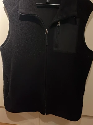 Svart fleeceväst med dragkedja - Mysig svart fleeceväst med hel dragkedja framtill och hög krage. Västen har en bröstficka med dragkedja och två sidofickor. Perfekt lager för kyliga dagar och enkel att matcha med olika outfits.