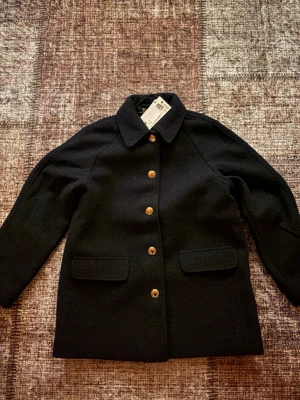 mörkblå ullkappa med guldknappar - Stilren mörkblå kappa i ull med klassisk krage och raka ärmar. Kappan har snygga guldknappar framtill och två fickor med lock. Aldrig använt! pris kan diskuteras.