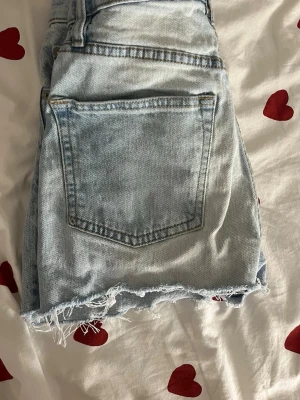 Ljusblå jeansshorts med fransig kant - Säljer ett par ljusblå jeansshorts från 157 med fransig, rå kant och slitna detaljer. Klassisk femficksmodell med knapp och dragkedja framtill. Perfekta för varma dagar och ger en avslappnad look.
