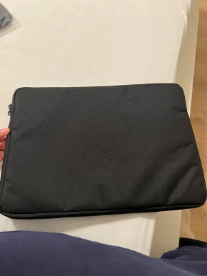 Svart laptopfodral i tyg - Snyggt och stilrent svart laptopfodral i mjukt tygmaterial. Fodralet har en enkel, rektangulär form och stängs med dragkedja längs kanten. Perfekt för att skydda din dator på språng och passar till de flesta laptops.