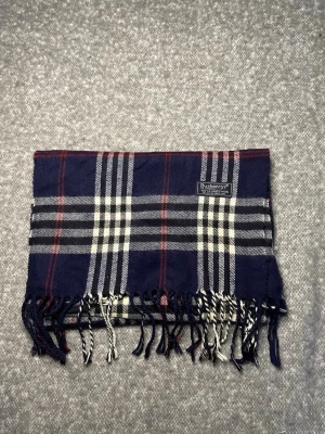 Burberry Halsduk Mörkblå - Klassisk Burberry-halsduk i 100 % lammull. Mjuk och varm med tidlöst rutmönster i mörkblå, svart, vitt och rött. Originaletikett, Made in England. 2MB