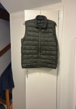 Olivgrön dunväst från Barbour - Snygg olivgrön dunväst från Barbour med quiltad design och hög krage. Västen har dragkedja framtill och diskret Barbour-logga på bröstet. Perfekt att slänga över en hoodie eller tröja för extra värme. Tillverkad i lätt och smidigt material.