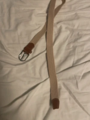 Beige flätat bälte med läderdetaljer - Snyggt beige flätat bälte med bruna läderdetaljer vid spännet och änden. Spännet är i silverfärgad metall och bältet har en stilren, enkel design som passar till många olika outfits.