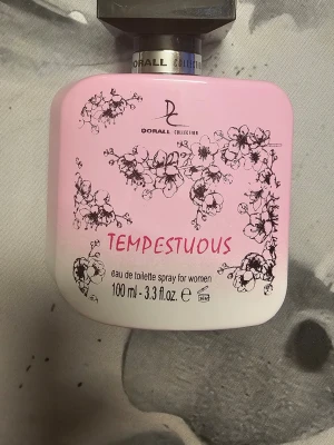  Dorall Tempestuous Eau de Toilette - En söt och feminin parfym från Dorall Collection i en rosa och vit flaska med blommigt mönster. Doften har fruktiga toppnoter, fresia i hjärtat och en varm bas av vaniljextrakt. Flaskan rymmer 100 ml och har en modern, stilren design.