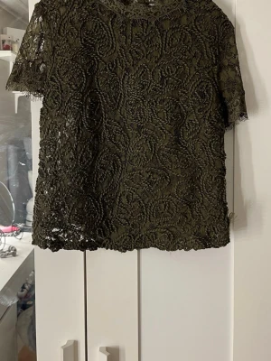 Olivgrön spetsblus från Zara - Snygg olivgrön topp från Zara Trafaluc i genombruten spets med blommigt mönster. Kortärmad modell med rund hals och råa kanter. Perfekt att styla lager på lager eller med ett linne under för en cool look.