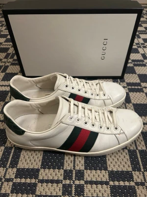 Gucci vita sneakers med röd/grön rand - Snygga vita sneakers från Gucci med klassiska rödgröna ränder på sidorna och gröna detaljer vid hälen. Skorna har snörning och är tillverkade i skinn för en lyxig känsla. Perfekta för dig som vill ha en ikonisk och stilren look.