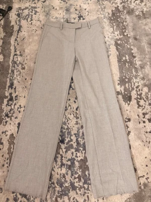 Beige kostymbyxa med raka ben - Stilrena beige kostymbyxor med raka ben och pressveck. Byxorna har hällor för skärp och klassisk midja. Perfekta för en clean och snygg look. Materialet känns lätt och har en diskret struktur.