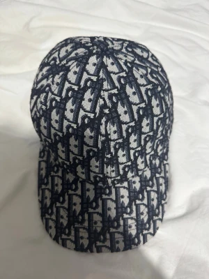 Dior keps med monogrammönster - Snygg keps från Dior med klassiskt blått och vitt monogrammönster över hela kepsen. Kepsen har böjd skärm och är tillverkad i ett tygmaterial med tydlig struktur. Perfekt accessoar för att lyfta din streetwear-look.