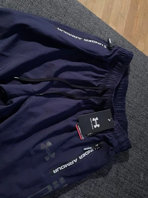 Mörkblå mjukisbyxor från Under Armour - Snygga mörkblå mjukisbyxor från Under Armour med resår i midjan och svarta dragsnören. Byxorna har dragkedjeförsedda fickor med logga och tryckt Under Armour-text längs benet. Perfekta för chill eller träning. Dom är lite väl små för att vara M så dom passar absolut i S. 