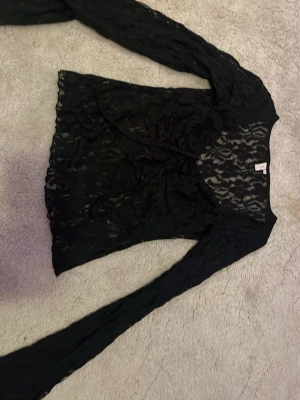 Svart spetsblus med lång ärm - Snygg svart blus i spets med långärmad design och omlott framtill. Transparent material som ger en cool och trendig look. Perfekt att styla med ett linne under eller som den är för en edgy vibe.
