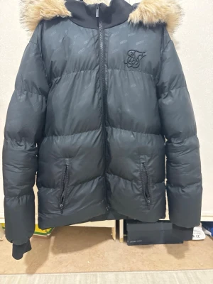 Svart pufferjacka med fuskpäls sikskilk - Säljer en svart pufferjacka från SikSilk med avtagbar huva och fuskpäls. Jackan har två dragkedjeförsedda fickor, ribbade muddar och diskret logga på bröstet. Perfekt för kalla vinterdagar och riktigt snygg streetstil.