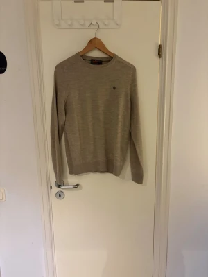 Beige stickad tröja från Morris  - Klassisk beige stickad tröja från Morris med rund halsringning och diskret logga broderad på bröstet. Tröjan har långa ärmar och ribbade muddar vid ärmslut och nederkant. Perfekt för lager-på-lager och enkel att matcha.