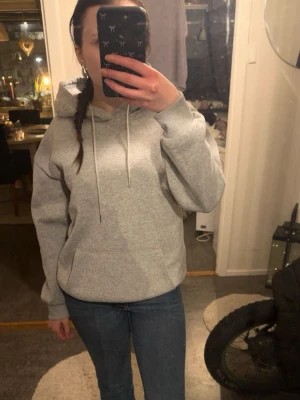 Grå basic hoodie med huva - Enkel och stilren grå hoodie med huva och dragsnören. Hoodien har en stor magficka och ribbade muddar vid ärmslut och nederkant. Perfekt för en avslappnad och bekväm look. Materialet känns mjukt och skönt mot huden.