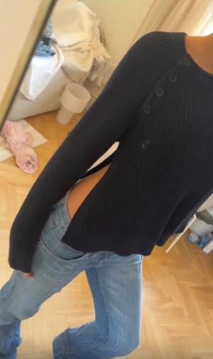 Mörkblå stickad tröja med knappar - Trendig mörkblå stickad tröja med långa ärmar och coola knappar längs axeln. Tröjan har en unik sidodetalj med öppen slits som ger en edgy look. Perfekt att styla med jeans för en avslappnad vibe.